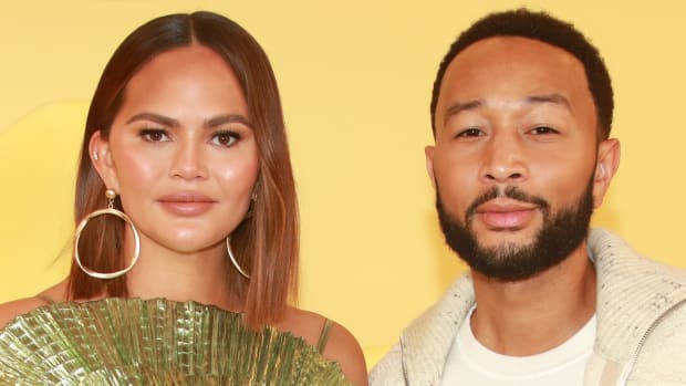 Chrissy Teigen and John Legend pose side-by-side in front of a yellow backdrop. Teigen wears a green pleated dress and Legend wears a white tee and cream-colored sweater.