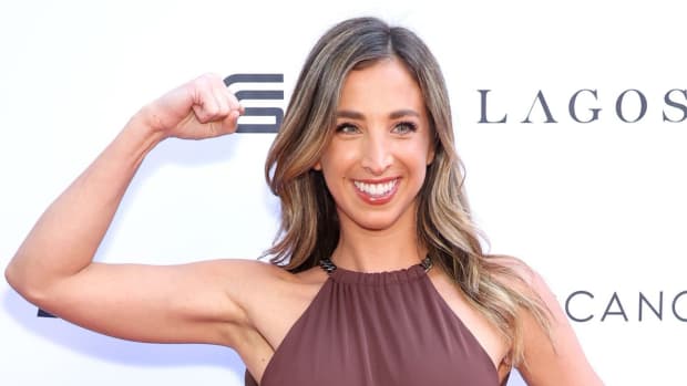 Katie Austin poses in a brown high-neck dress and flexes her arm muscle.