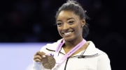 Simone Biles smiles for the camera in her white Nike zip-up jacket and her Worlds Championship medal.