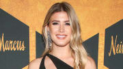 Genie Bouchard poses in front of a gold and black backdrop, wearing a black one-shoulder dress and her blonde hair in a soft wave.