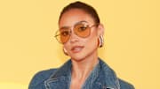 Shay Mitchell poses in a denim jacket, oversized gold earrings and orange-tinted sunglasses.