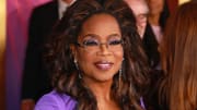 Oprah Winfrey poses in a bright purple dress and round wire-rimmed glasses and smiles for the camera.
