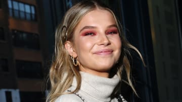 Ellie Thumann wears a gray turtle neck and hot pink eye makeup smiles at the camera.