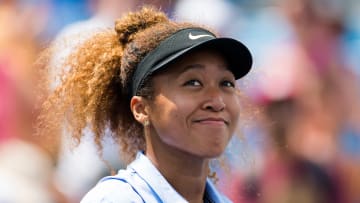 Naomi Osaka wears a blue collared shirt and a black Nike visor and smiles at the crowd before a tennis match.