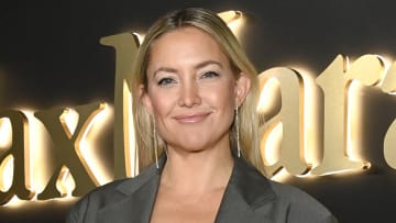 Kate Hudson poses in a gray blazer and silver drop earrings and smiles for the camera.