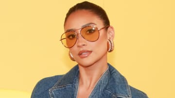 Shay Mitchell poses in a denim jacket, oversized gold earrings and orange-tinted sunglasses.