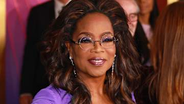 Oprah Winfrey poses in a bright purple dress and round wire-rimmed glasses and smiles for the camera.