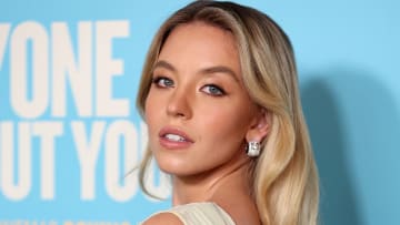 Sydney Sweeney poses in a cream mesh dress and diamond huggie earrings and looks over her shoulder at the camera.