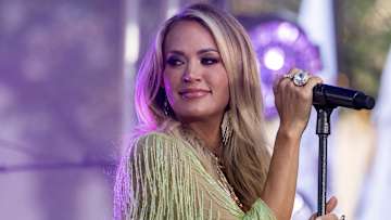Carrie Underwood poses in front of a microphone in a green fringed top and smiles.