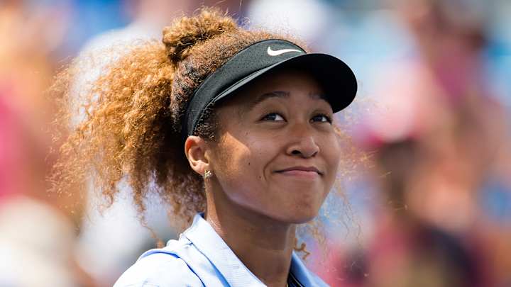 Naomi Osaka wears a blue collared shirt and a black Nike visor and smiles at the crowd before a tennis match.
