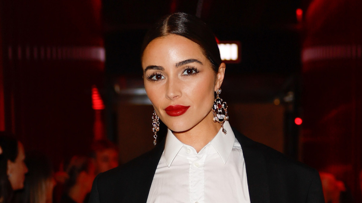 Olivia Culpo sports a slicked-back up-do, a red lip and oversized silver dangle earrings and smiles at the camera.