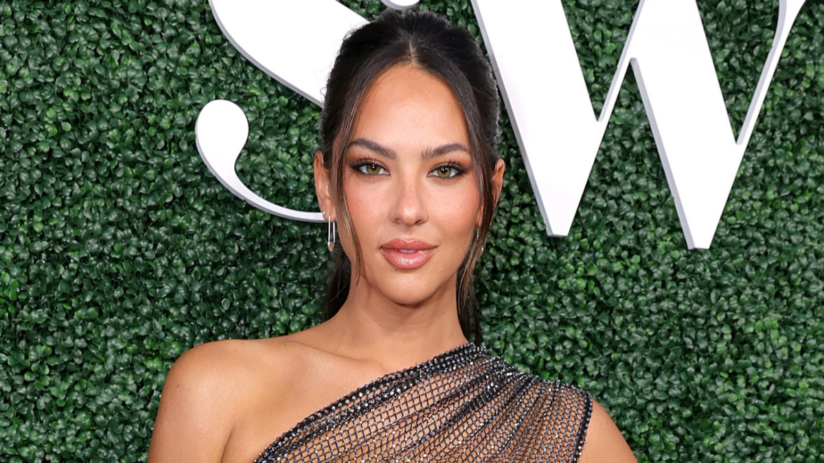 Christen Harper poses in front of a green backdrop in a black mesh one-shoulder dress.