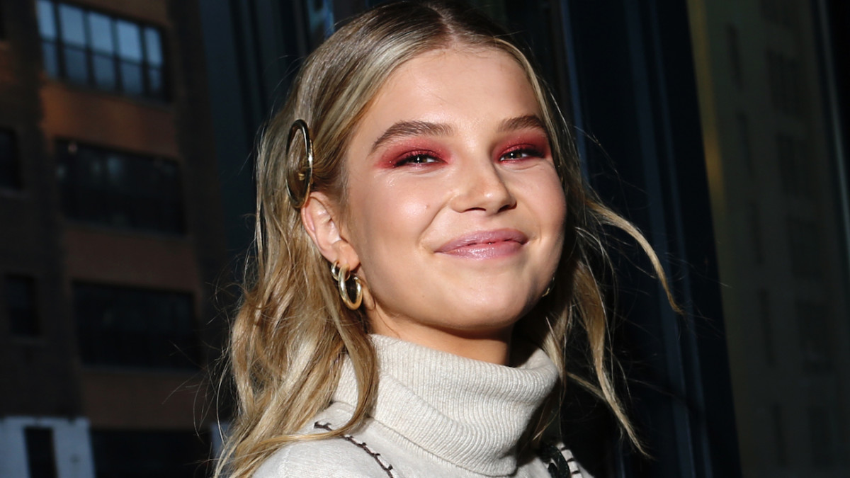 Ellie Thumann wears a gray turtle neck and hot pink eye makeup smiles at the camera.