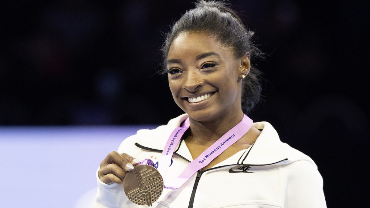 Simone Biles smiles for the camera in her white Nike zip-up jacket and her Worlds Championship medal.