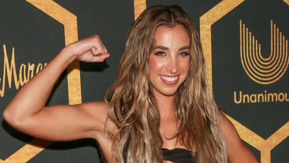 Katie Austin flexes her arm muscle and smiles at the camera wearing a black dress and her dirty blonde hair in a beach wave.