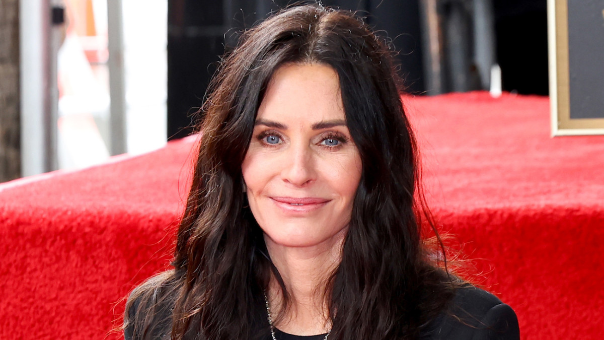 Courteney Cox smiles at the camera wearing a black blazer and her dark brown hair down in a loose wave.