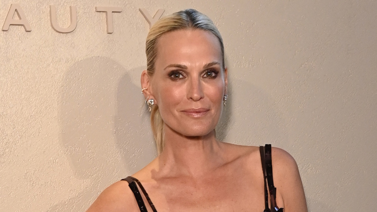 Molly Sims smiles in a black dress in front of a cream-colored backdrop.