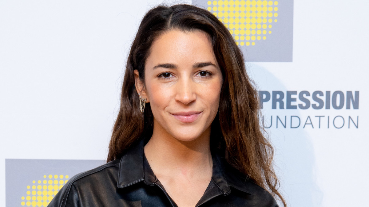 Aly Raisman poses in a black leather top and silver chain earrings and smiles softly for the camera.