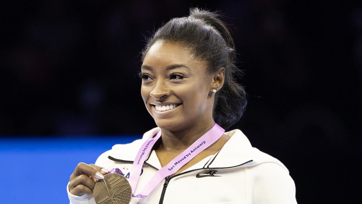 Simone Biles poses in a white quarter zip and sports a gymnastics medal around her neck.
