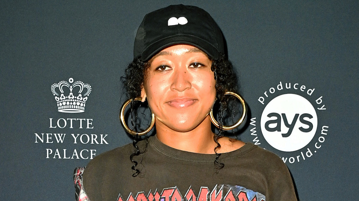 Naomi Osaka poses in a graphic tee, black ball cap and oversized hoops and smiles for the camera.