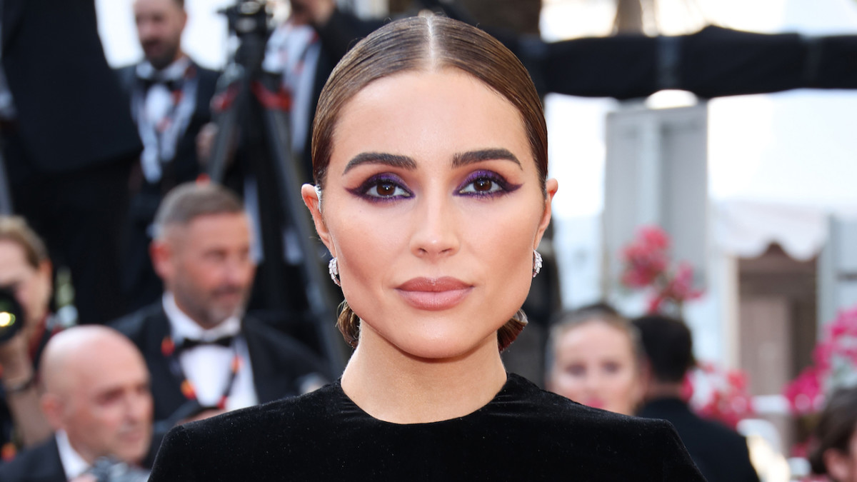 Olivia Culpo poses in a black velour top, diamond earrings and edgy eye makeup and smiles softly for the camera.